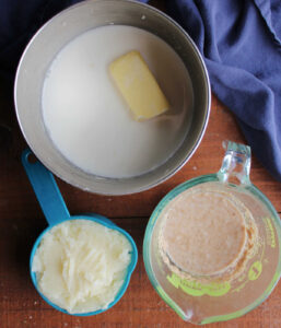 Milk, butter, and sugar in saucepan next to a cup of mashed potatoes and a measuring cup with yeast mixture in it.