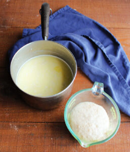 Saucepan with potato, milk, sugar mixture next to measuring cup with foamy yeast mixture inside.
