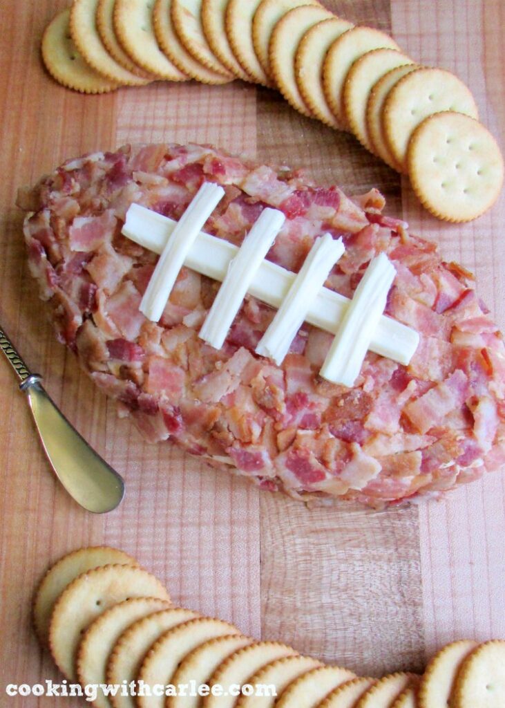 Football shaped bacon coated pig skin cheese ball on maple cutting board with crackers nearby.