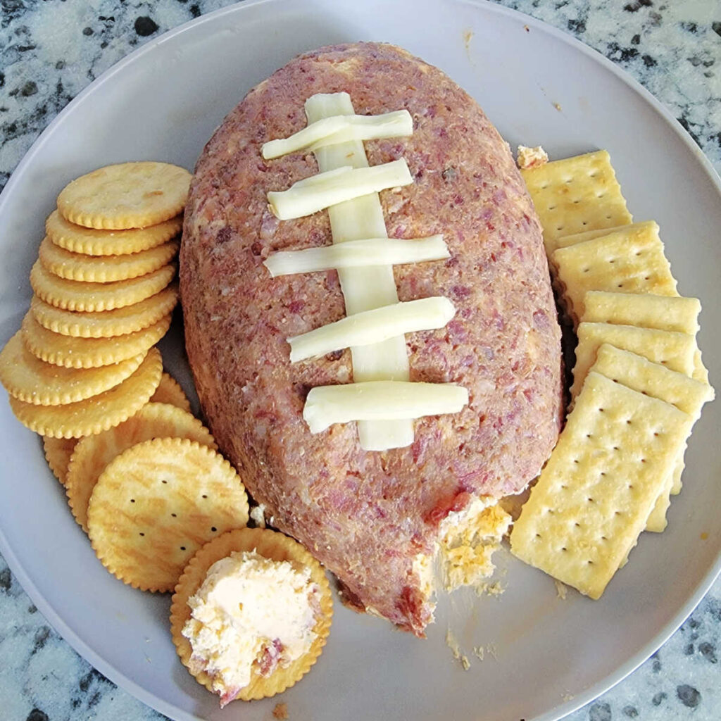 Plate of cheddar bacon ranch cheese ball in the shape of a football with crackers.