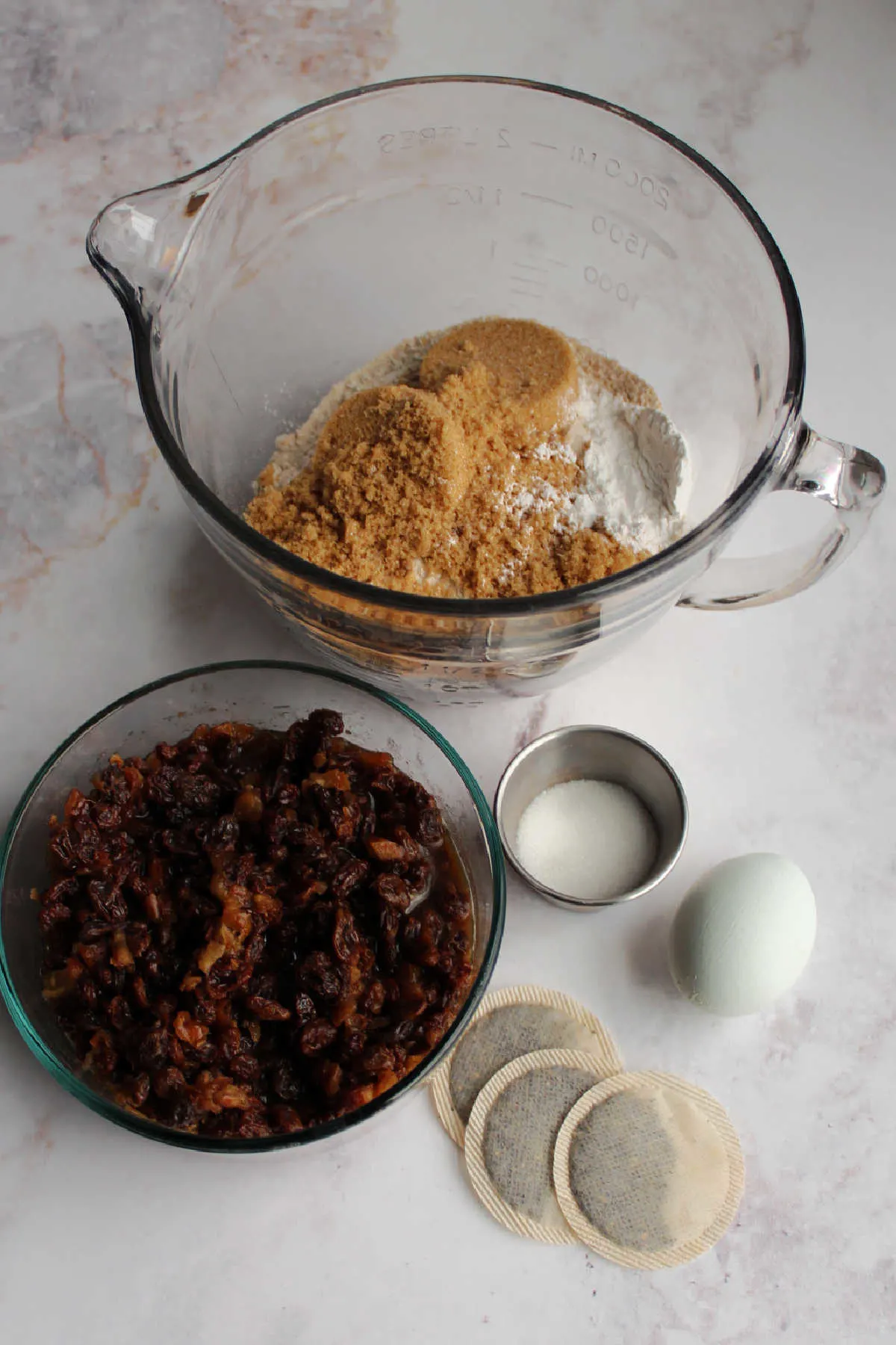 Irish tea brack ingredients including bowl of flour and sugar, raisins and dates soaking in tea, tea bags, sugar and an egg ready to be made into batter.