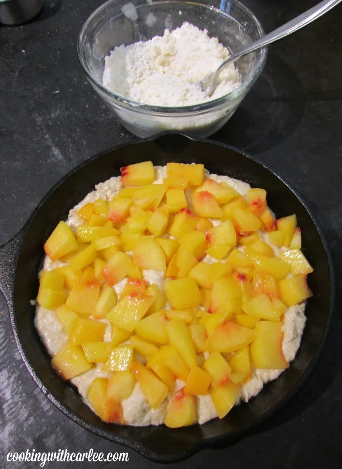 chopped peaches spread over cake batter in skillet with crumb topping in bowl nearby.