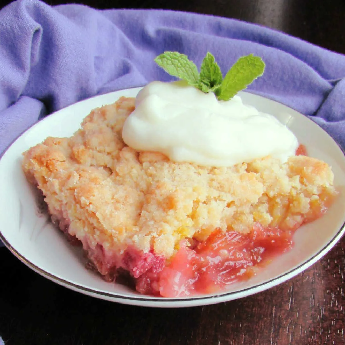 Small bowl with piece of rhubarb crunch showing pink layer topped with golden buttery crumb topping with a dollop of yogurt and sprig of mint on top.