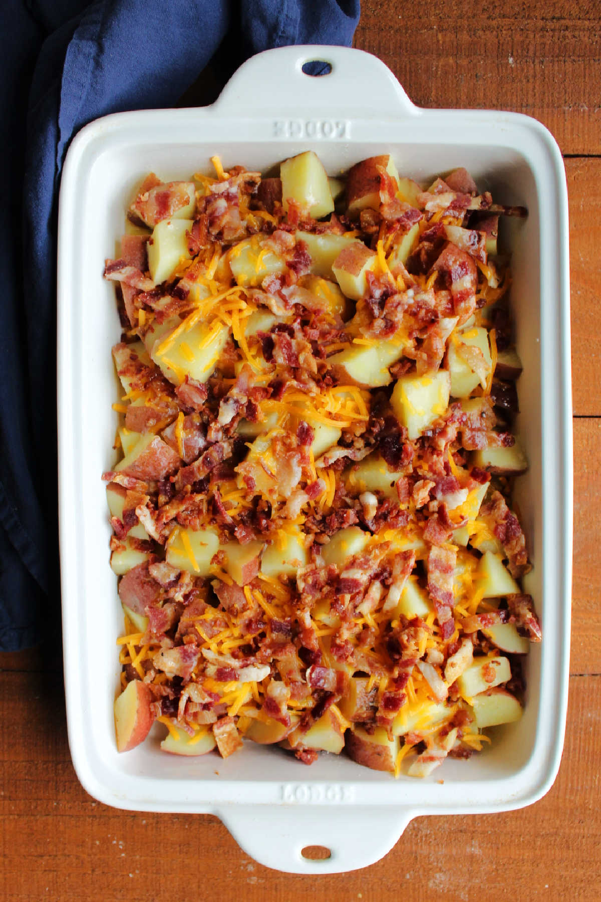 Casserole dish with cooked potatoes topped with shredded cheddar cheese and chopped cooked bacon ready to go in the oven.