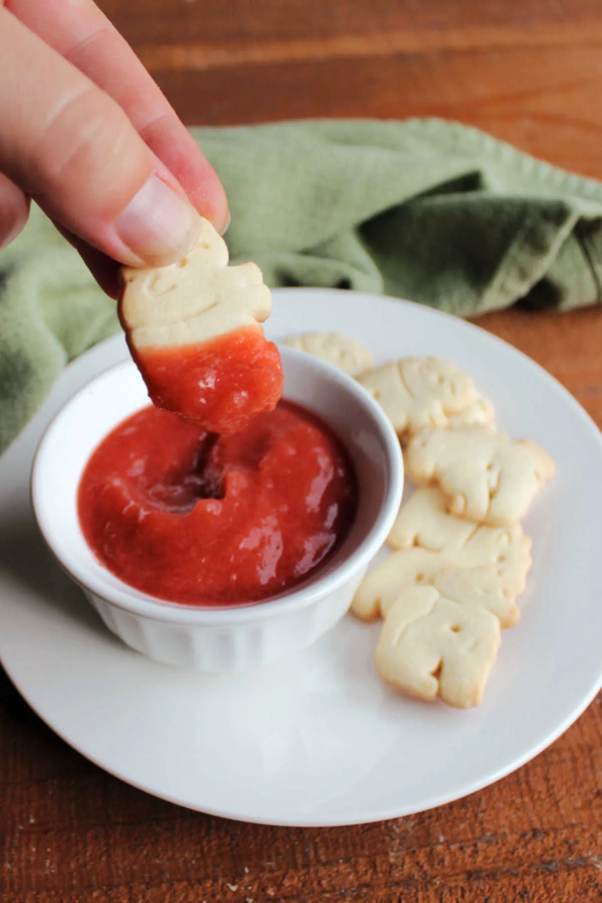 Hand dipping an animal cracker into the bowl of pink strawberry rhubarb sauce showing it sticking to the cookie.