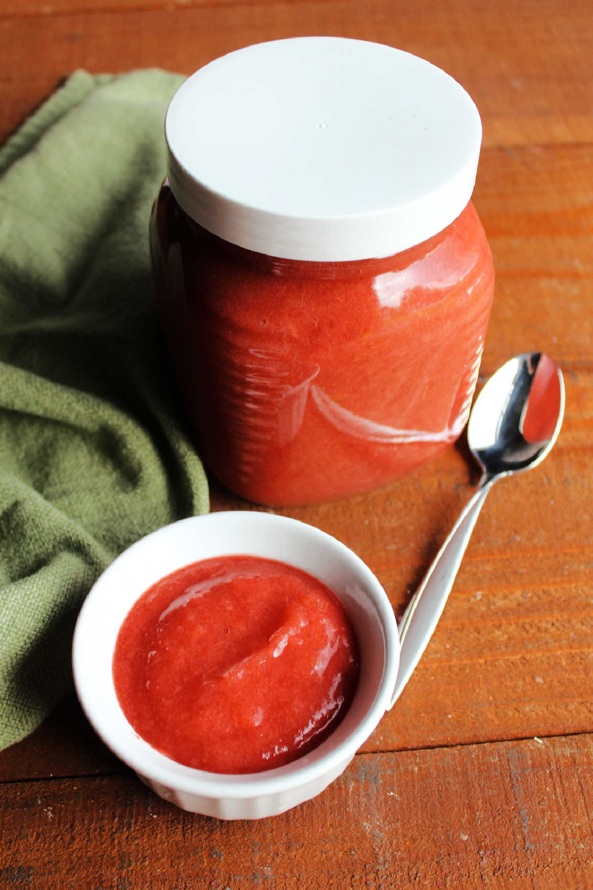 Small ramekin of pink strawberry rhubarb sauce with spoon next to large jar of strawberry rhubarb applesauce. 