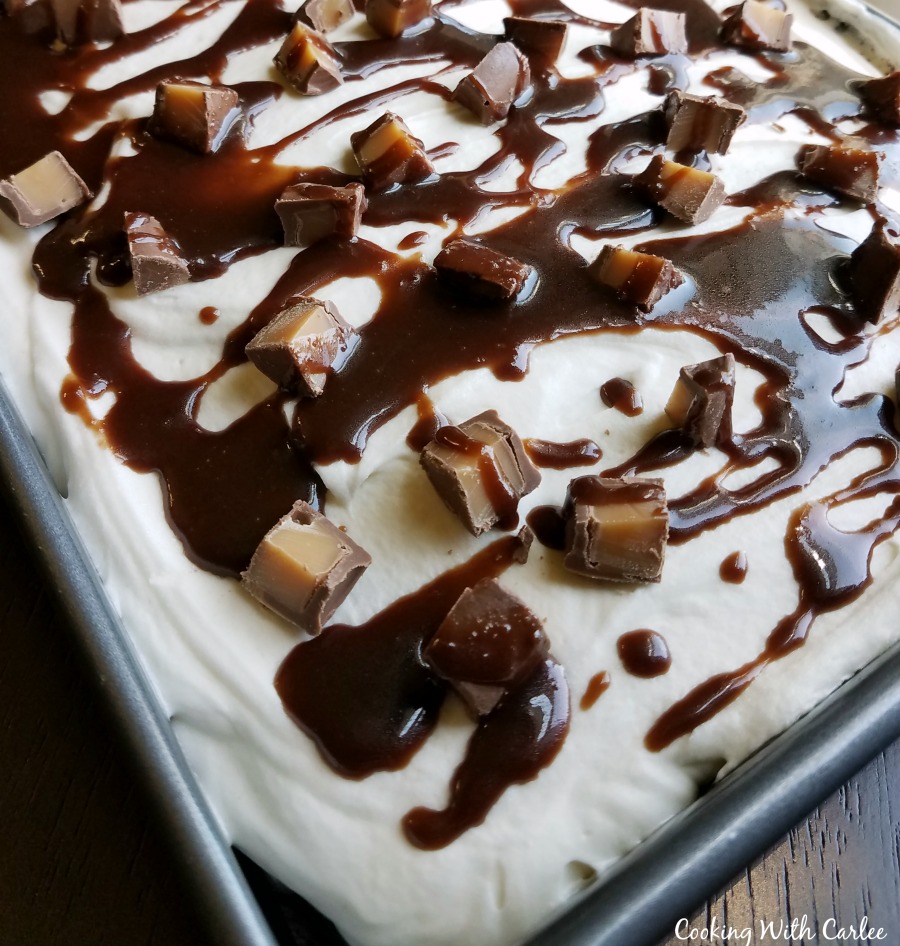 corner of cake in pan with white frosting, fudge drizzle and chopped candies showing.