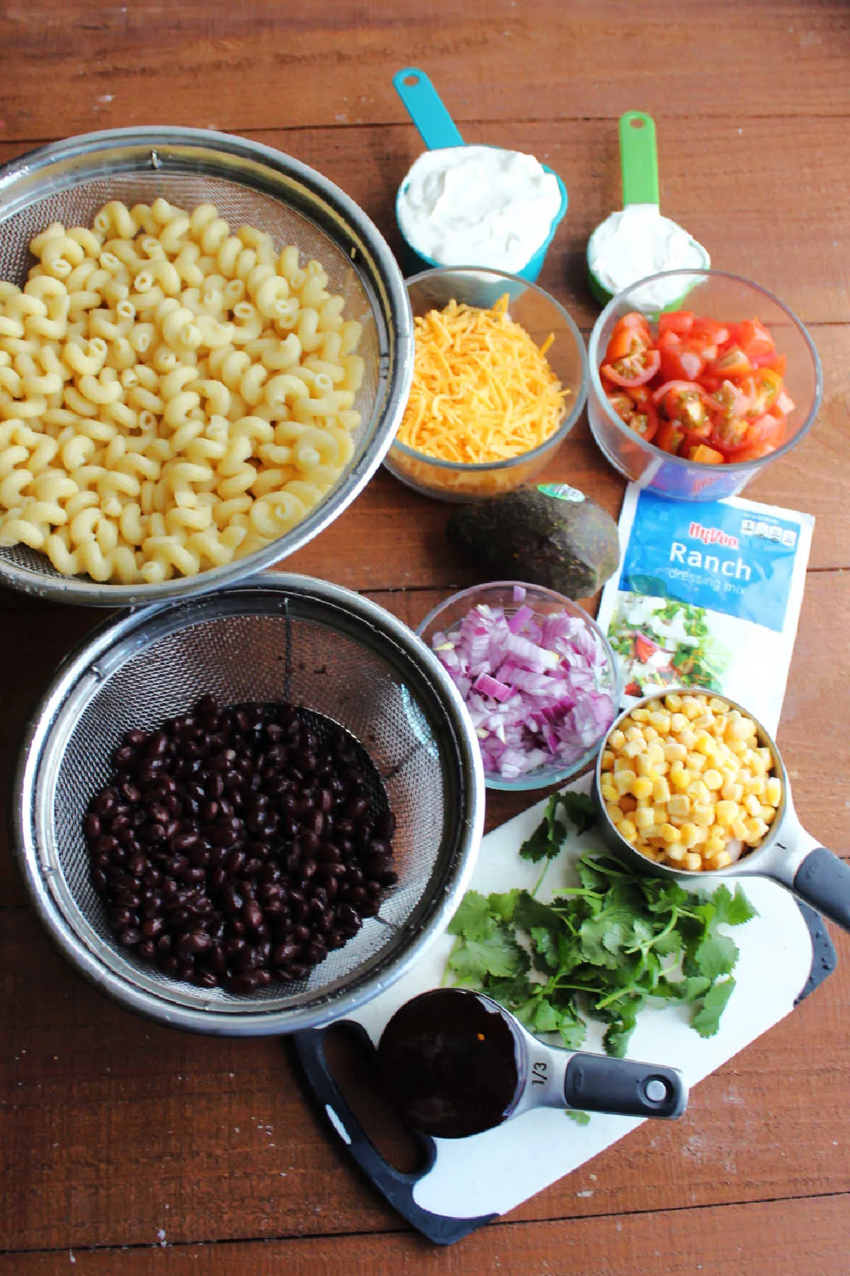 Ingredients including cooked pasta, cheese, sour cream, ranch seasoning, corn, black beans, red onion, bbq sauce and cilantro ready to be made into bbq ranch macaroni salad.