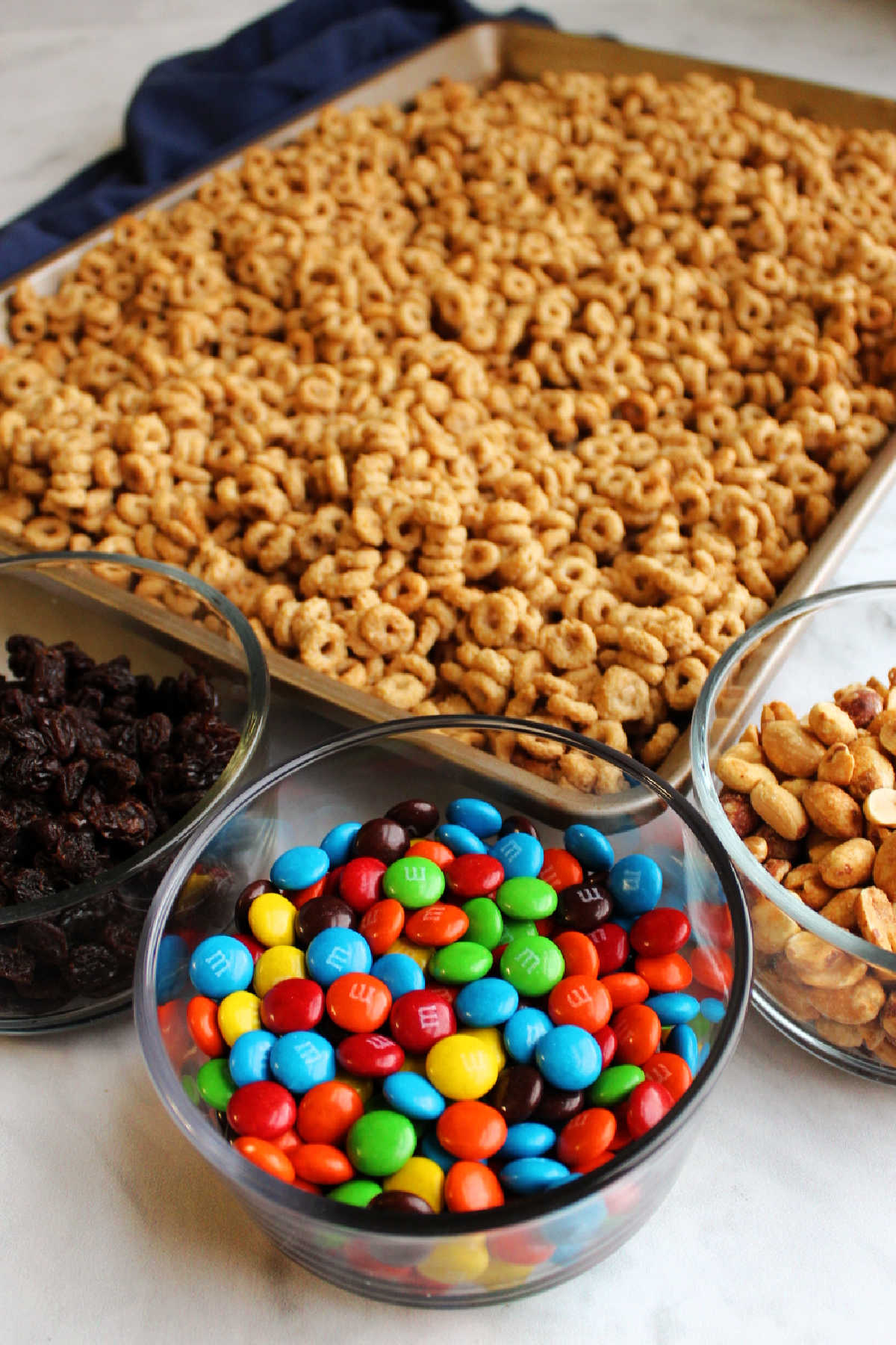 Sheet pan filled with maple peanut butter coated cheerios next to bowls of M&Ms, raisins and peanuts.
