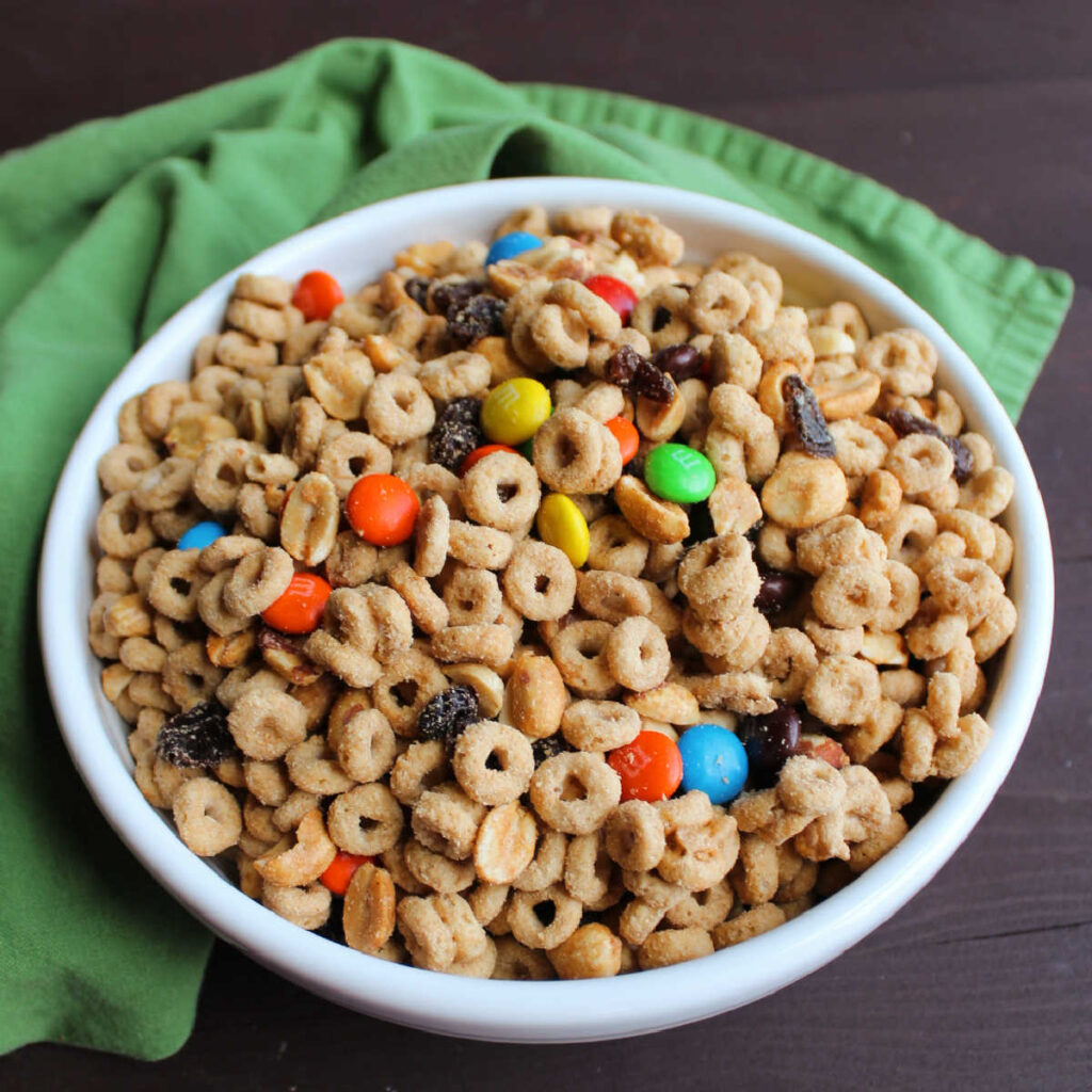 Serving bowl of gorp snack mix made with peanut butter and maple coated cheerios, raisins, peanuts and chocolate candies.