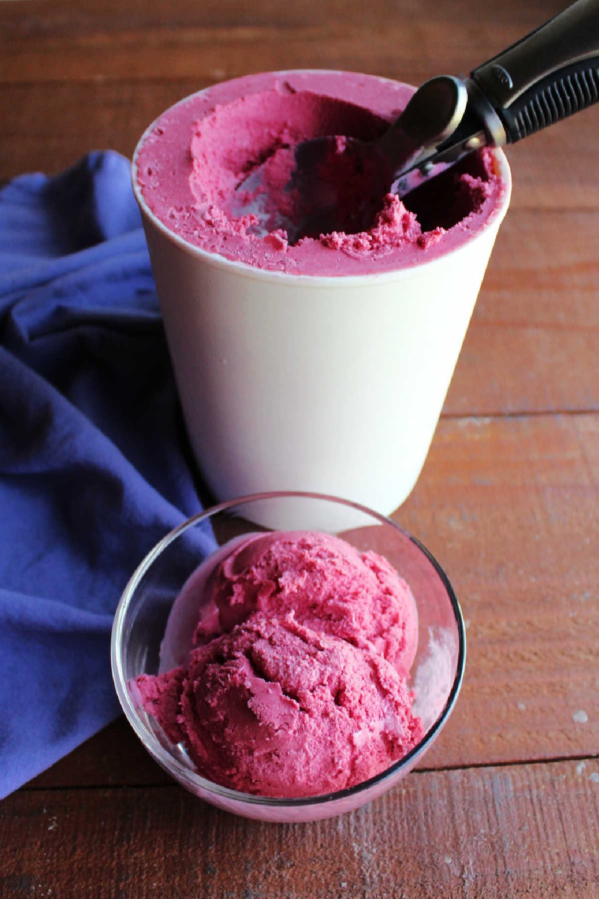 Scoops of vibrant blackberry ice cream in small glass bowl with larger container of homemade blackberry ice cream in the background.