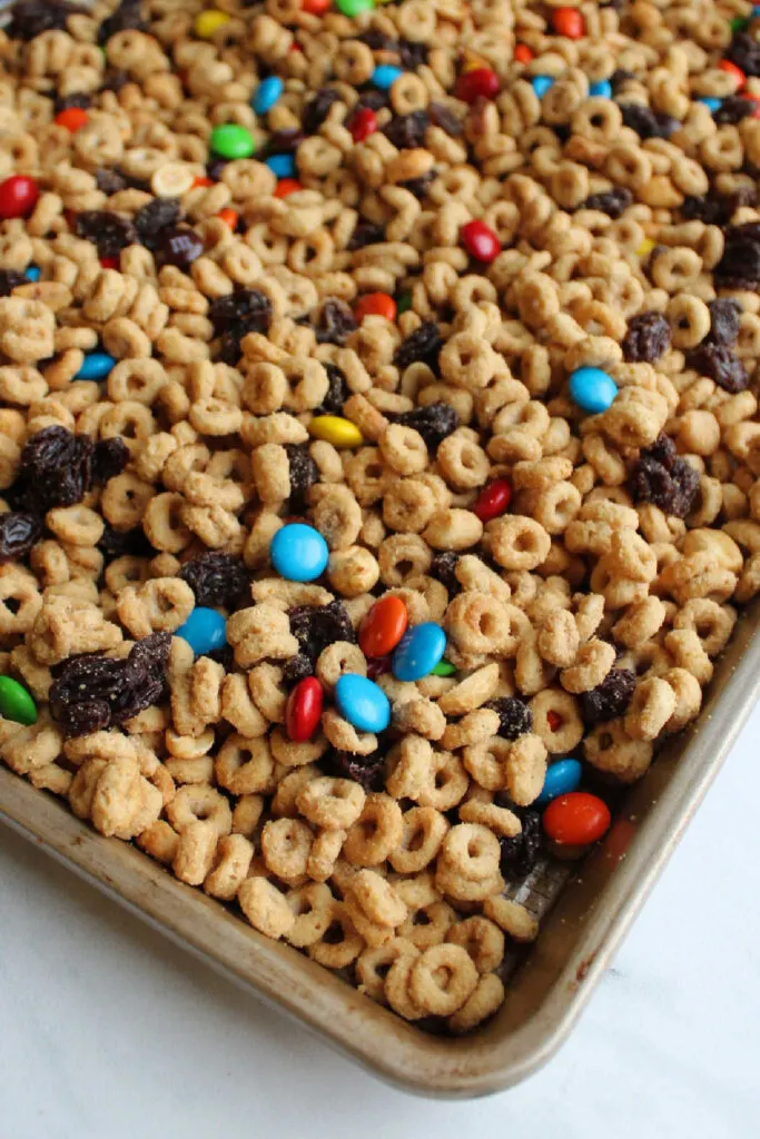 Sheet pan filled with trail mix style gorp snack mix with maple peanut butter coated cereal, candies, raisins and peanuts.