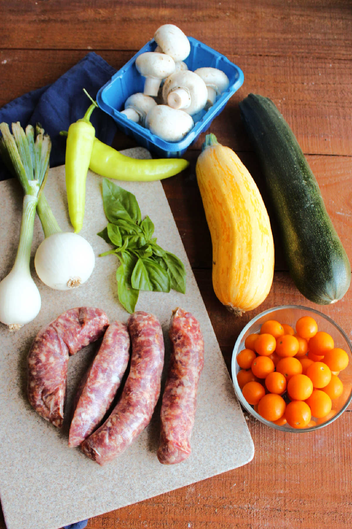 Ingredients including Italian sausage links, zucchini, yellow squash, cherry tomatoes, basil, spring onions, banana peppers and button mushrooms ready to be made into sheet pan meal.