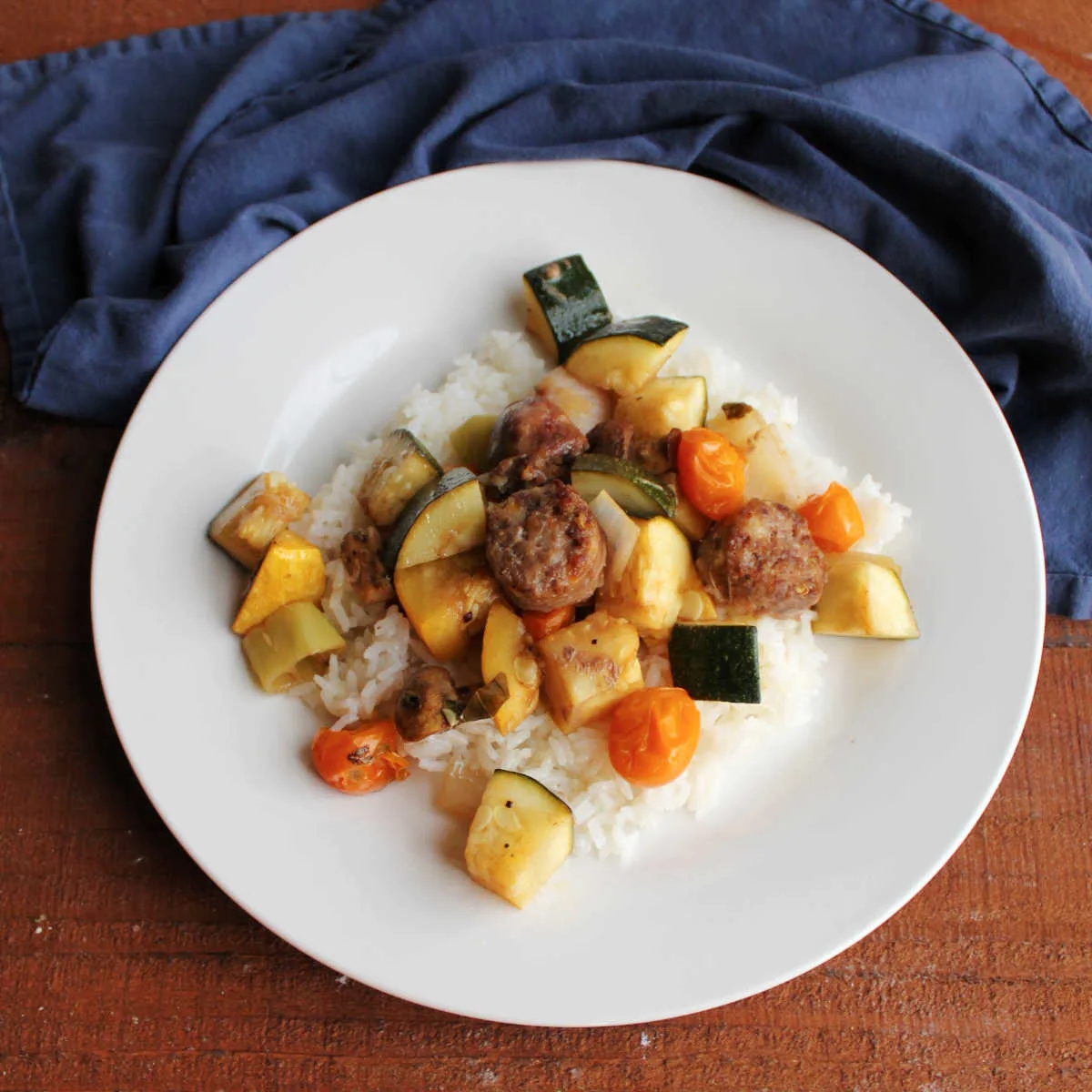 Dinner plate with rice under roasted Italian sausage chunks and vegetables including zucchini, yellow squash, cherry tomatoes, and more.