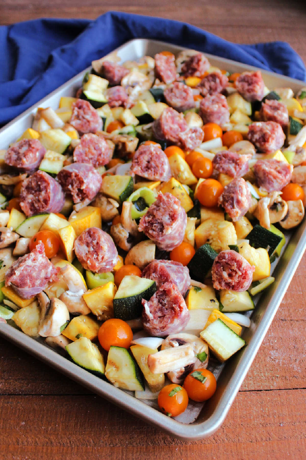 Vegetables on rimmed sheet pan topped with pieces of raw Italian sausage links, ready to be roasted.