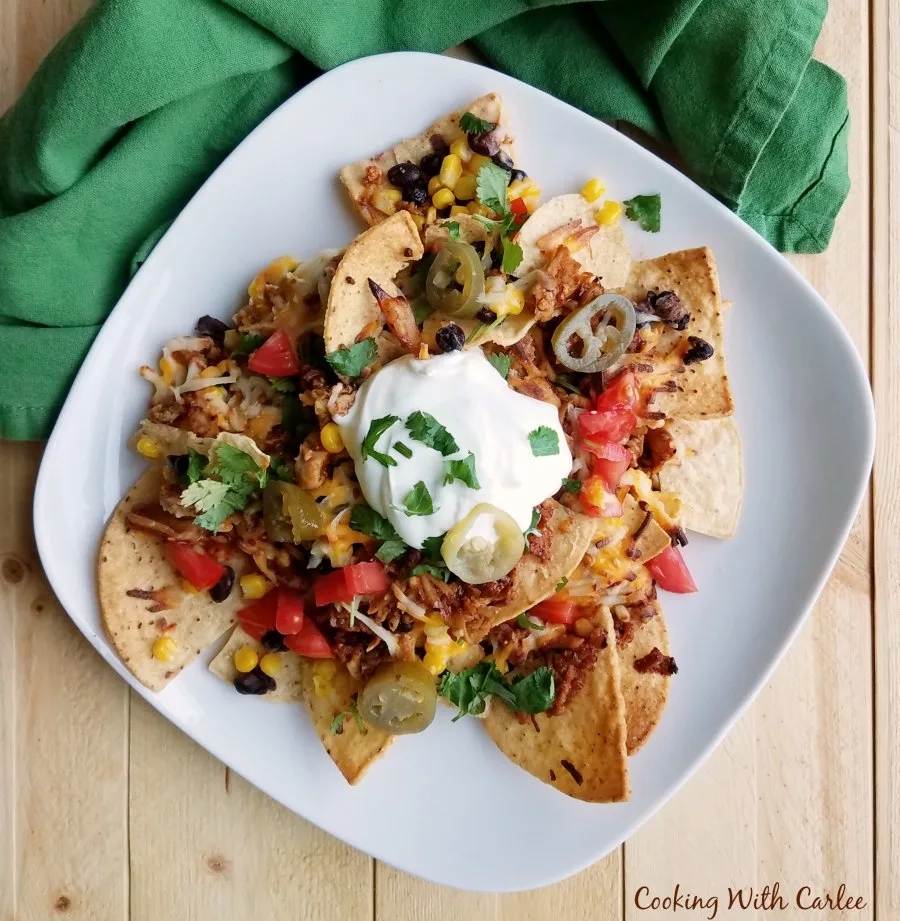 Plate piled with chips, pulled pork, veggies, and more for yummy pulled pork nachos topped with sour cream, pickled jalapenos, and chopped fresh cilantro.