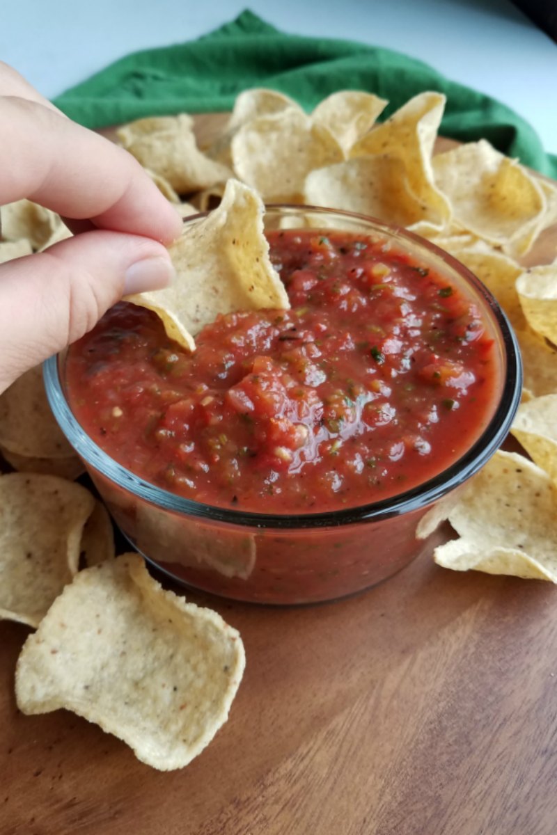 Hand dipping a tortilla scoop into a glass bowl of thick homemade salsa made from roasted garden fresh veggies.