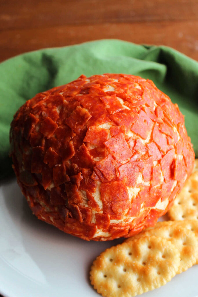 Pizza flavored cheese ball coated in pepperoni next to crackers ready to serve. 