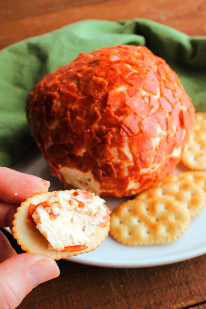 Hand holding cracker with pepperoni cheese ball spread over it.