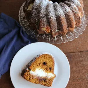 Slice of pumpkin bundt cake with cream cheese filling and chocolate chips on plate with remaining powdered sugar dusted cake in background.