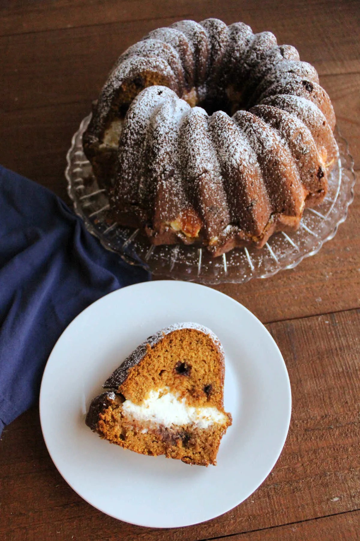 Slice of cream cheese stuffed pumpkin bundt cake with chocolate chips on plate with remaining powdered sugar dusted cake in the background.