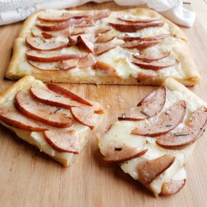 Pieces of pear and brie flatbread on cutting board showing gooey cheese, soft pear, and herbs.