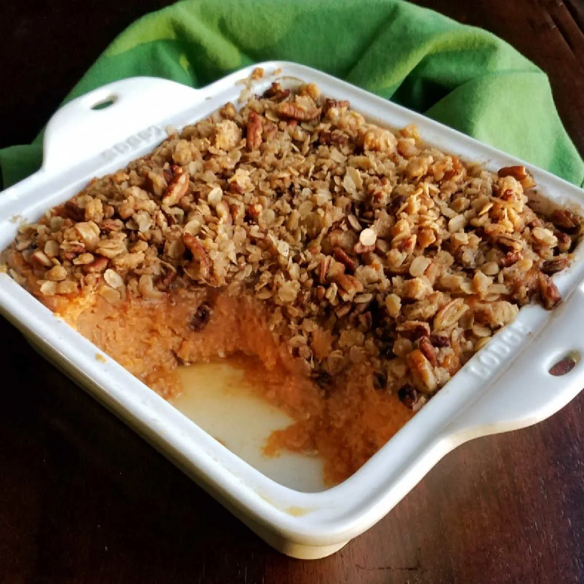 Square baking dish filled with oat and pecan topped casserole with some missing showing smooth sweet potato layer underneath.