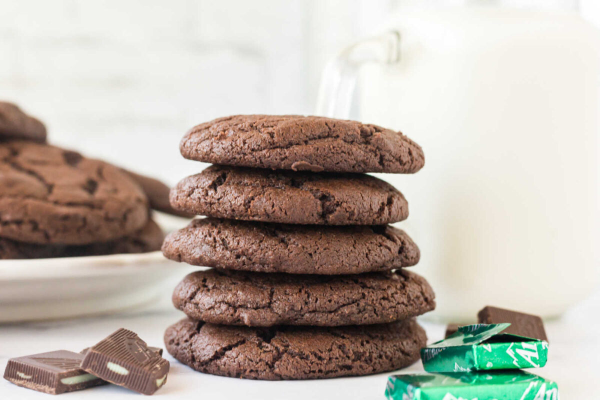 Stack of chocolate cookies with bits of Andes mints inside next to jug of milk and more chocolate mint candies.