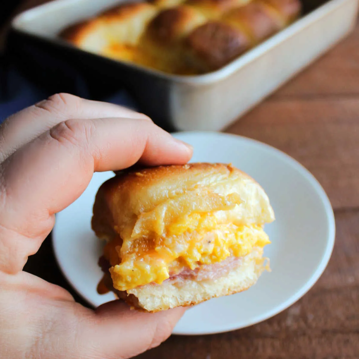 Close up of a hand holding a breakfast slider with sliced ham, scrambled eggs and melted cheese.