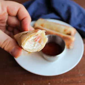 Hand holding half of a crab rangoon egg roll showing creamy filling with bits of pink crab meat inside.