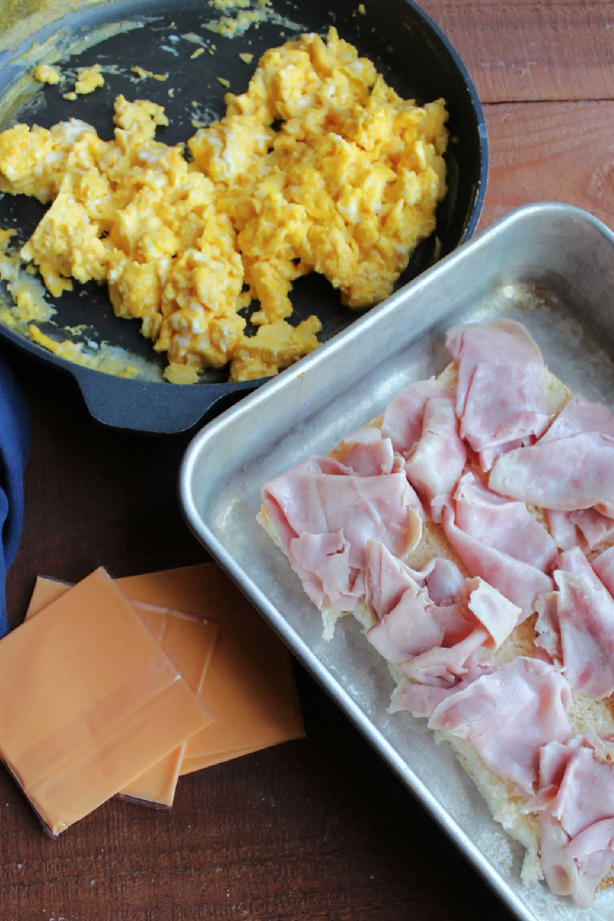 Skillet of scrambled eggs next to slices of cheese and bottom of dinner rolls topped with ham, ready to assemble breakfast sliders.