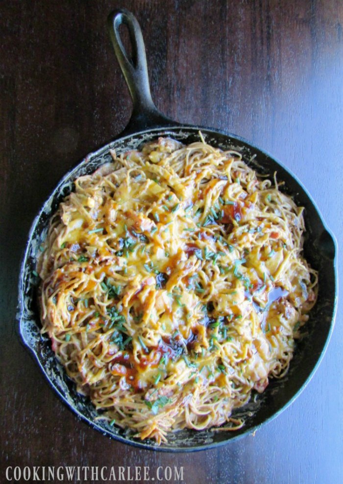 cast iron skillet full of bbq pulled pork spaghetti with melted cheese and herbs on top