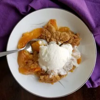 Serving of peach cobbler with warm peach slices and brown sugar biscuit like tipping served with a scoop of vanilla ice cream.