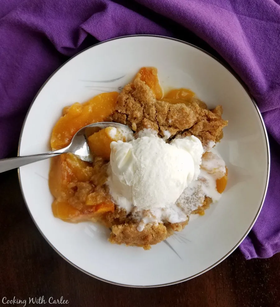 Serving of peach cobbler with warm peach slices and brown sugar biscuit like tipping served with a scoop of vanilla ice cream. 