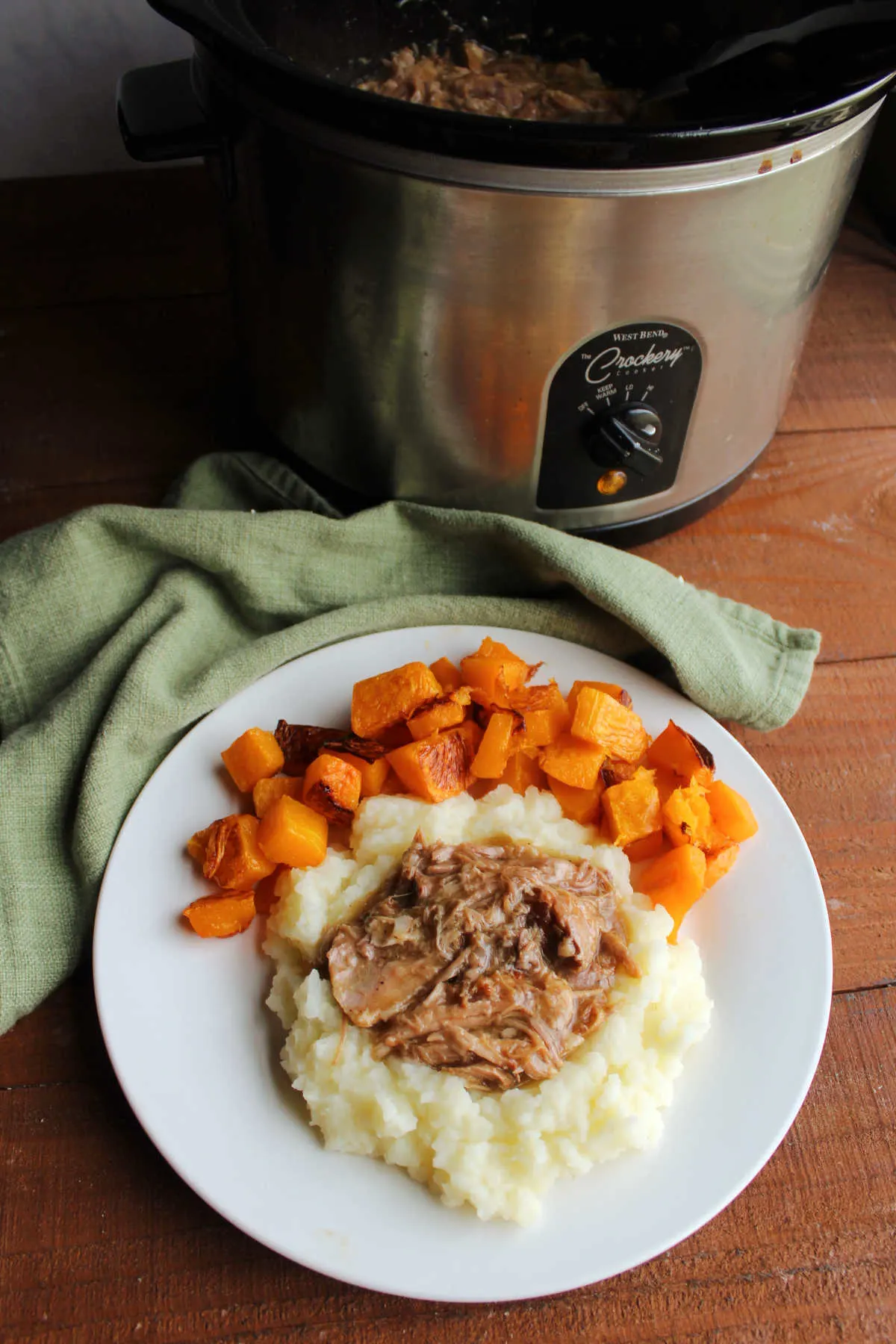 Dinner plate with apple cider pulled pork on potatoes with butternut squash next to crockpot with remaining pork mixture inside.