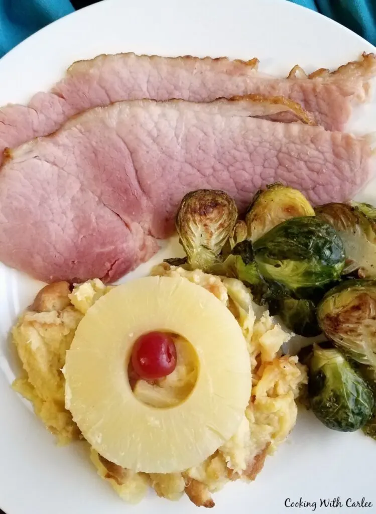 Close up of a dinner plate with scalloped pineapple, slices of ham, and roasted brussels sprouts.