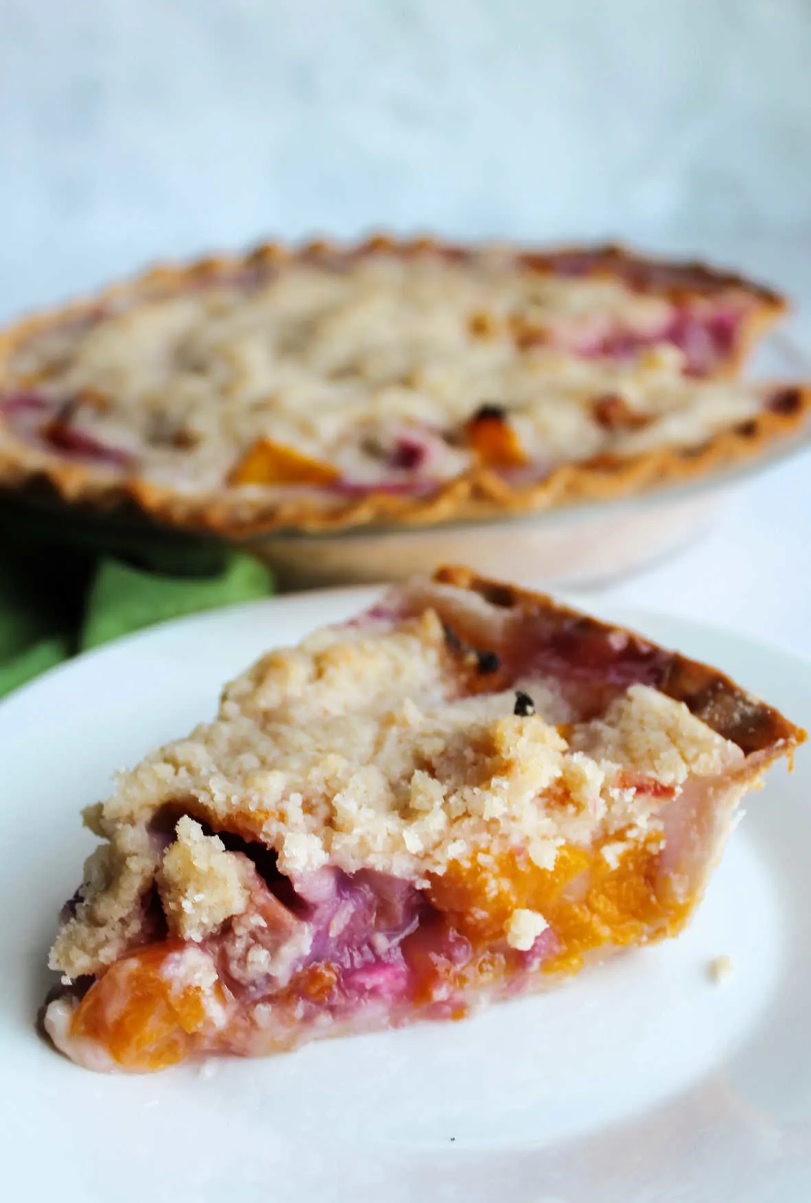 Slice of crumb topped peach rhubarb pie in front of remaining pie.