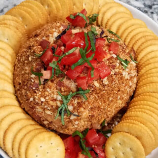 bacon bruschetta cheese ball topped with diced tomatoes and basil chiffonade with a ring of crackers around it.