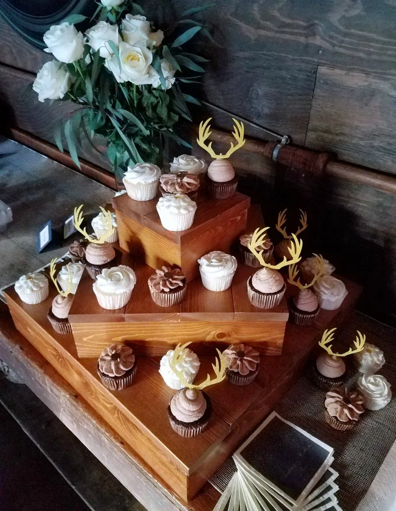 Wedding cupcakes arranged on wooden boxes, some with frosting flowers piped on top and some with gold glitter antler toppers.