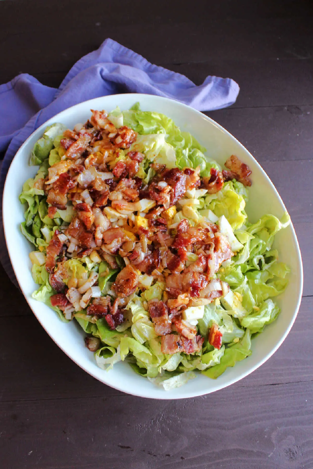 Bowl of lettuce topped with hard boiled eggs and warm bacon dressing ready to be tossed.