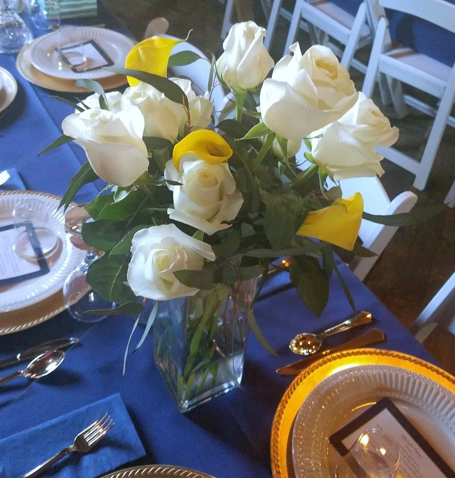 Bouquet of white roses and yellow calla lilies on table with navy tablecloth and gold and white place settings.
