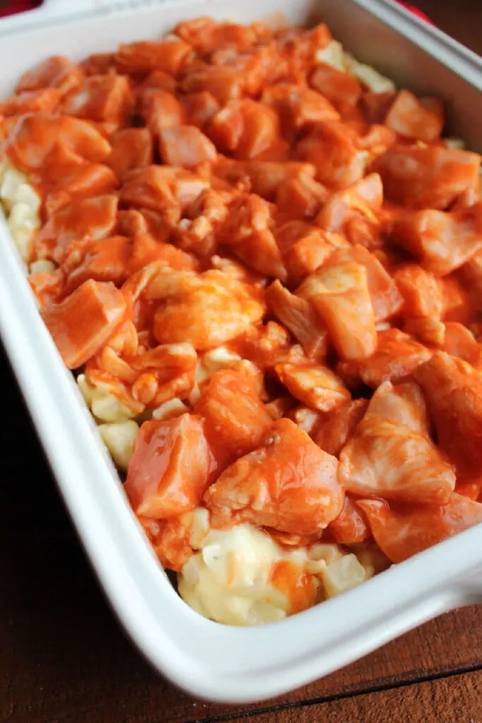 Raw chicken tossed in buffalo sauce and arranged over cheesy ranch potatoes in pan.