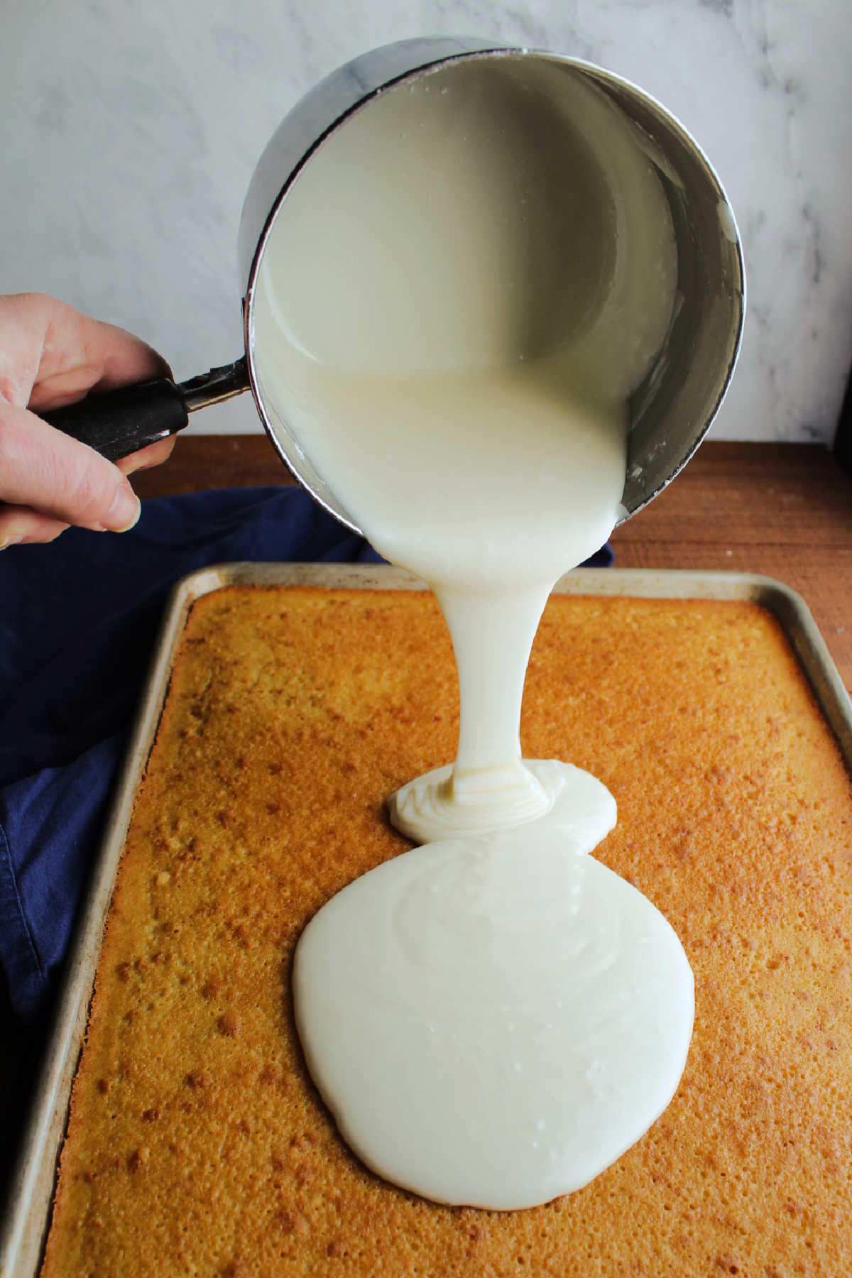 Pouring warm vanilla icing out of saucepan and onto baked vanilla Texas sheet cake.