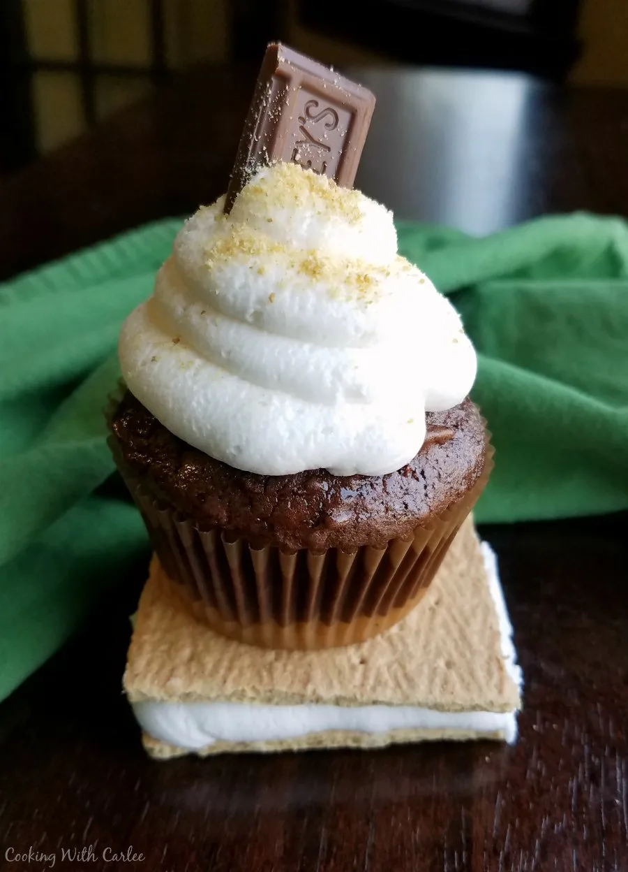 s'mores cupcake perched on top of a graham cracker and toasted marshmallow sandwich 