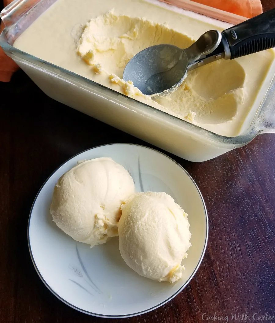 Two scoops of creamy orange ice cream in small bowl with glass loaf pan with more ice cream in the background. 