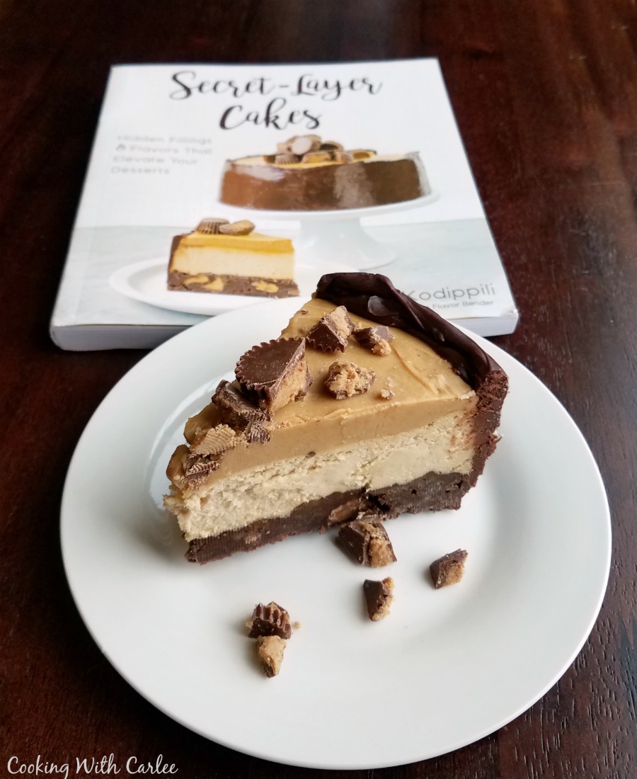 Slice of layered peanut butter and chocolate cheesecake with cookbook in background.