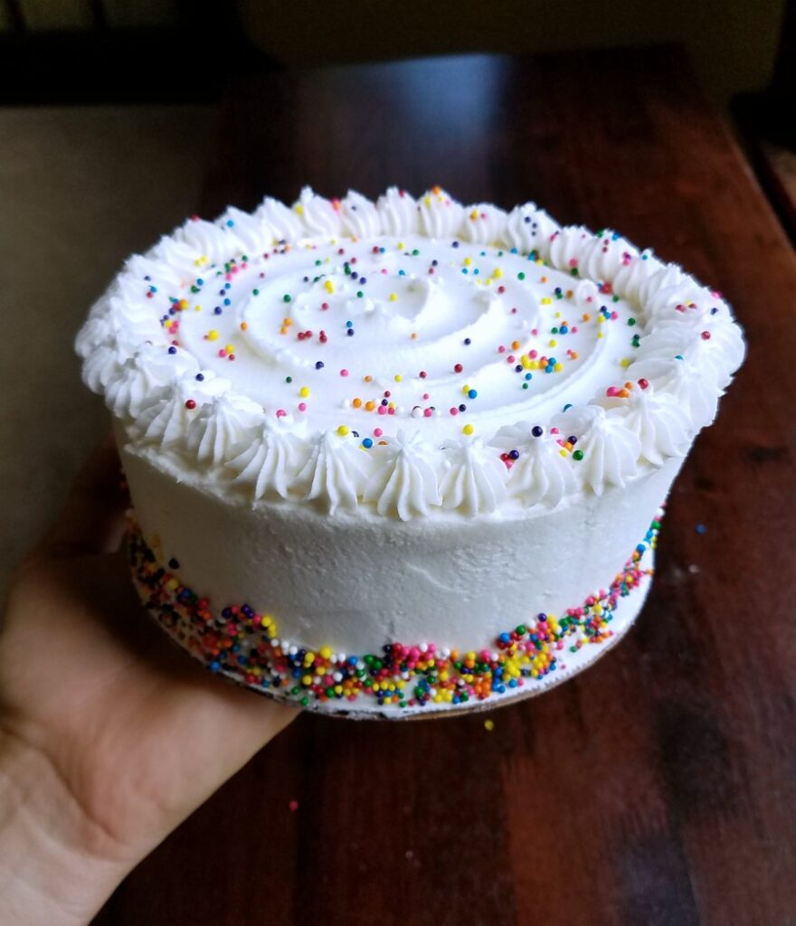 small round cake with white frosting, sprinkle decoration and piped top border.