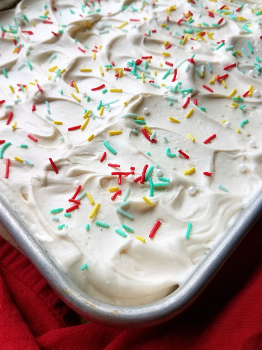 corner of sheet cake with fluffy white whipped sweetened condensed milk frosting made into light swirls with the back of a spoon topped with sprinkles. 