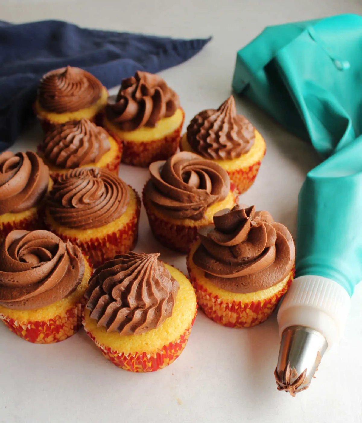 Piping bag filled with fudge frosting next to vanilla cupcakes topped with swirls of piped fudge frosting. 