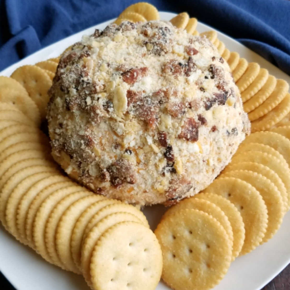 Cheddar bacon ranch cheese ball coated in cracker crumbs and bacon pieces surrounded by butter crackers on plate, ready to serve.
